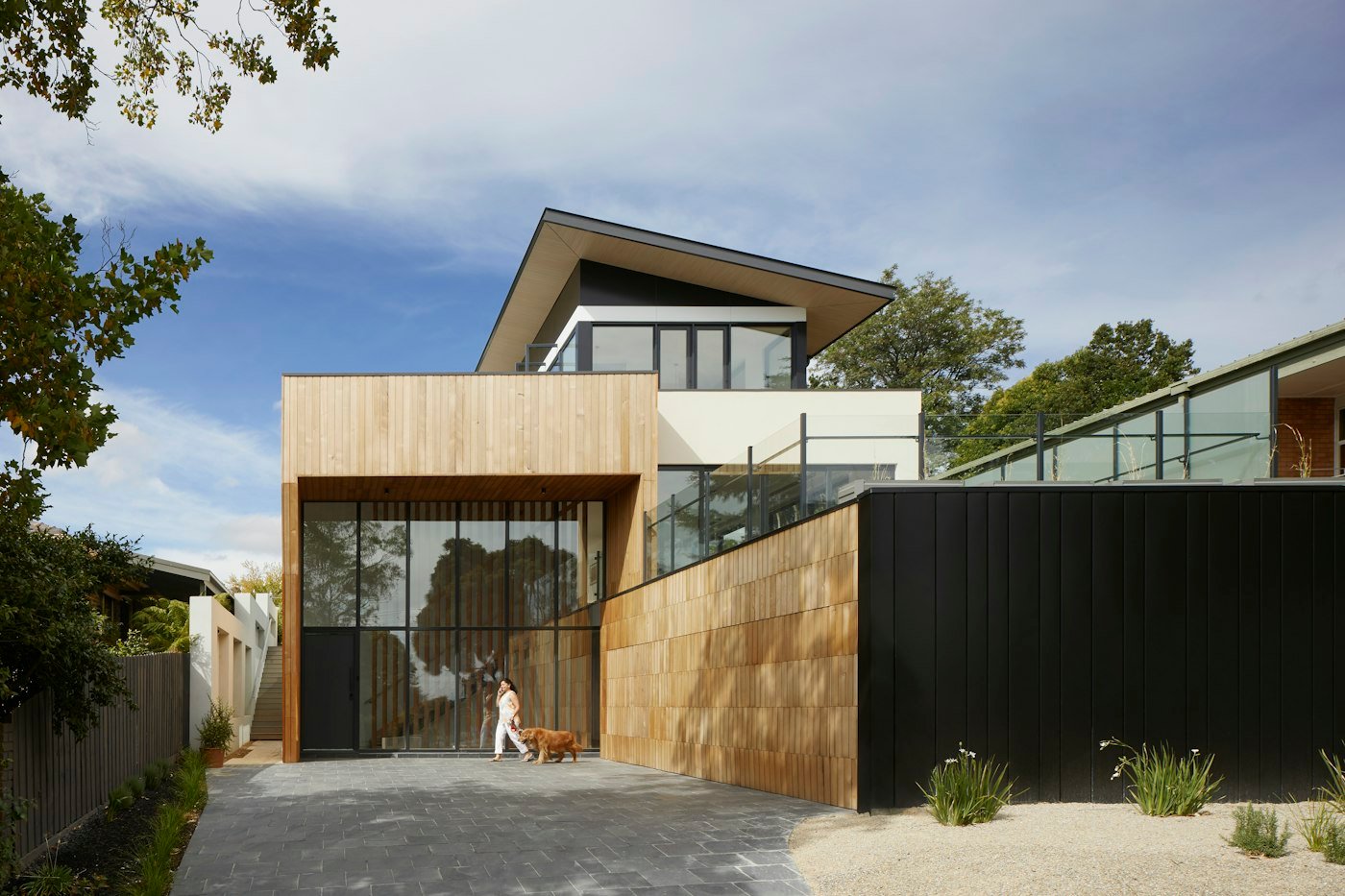 A timber and black modern house, with a person and a dog standing on the paved drive
